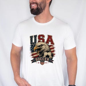 Puede incluir: Una camiseta blanca de manga corta con un estampado gr&aacute;fico de un &aacute;guila calva, un bal&oacute;n de f&uacute;tbol y la bandera estadounidense. El texto "USA" aparece en rojo y azul sobre el &aacute;guila, y la palabra "SOCCER" est&aacute; impresa en una banda azul debajo.