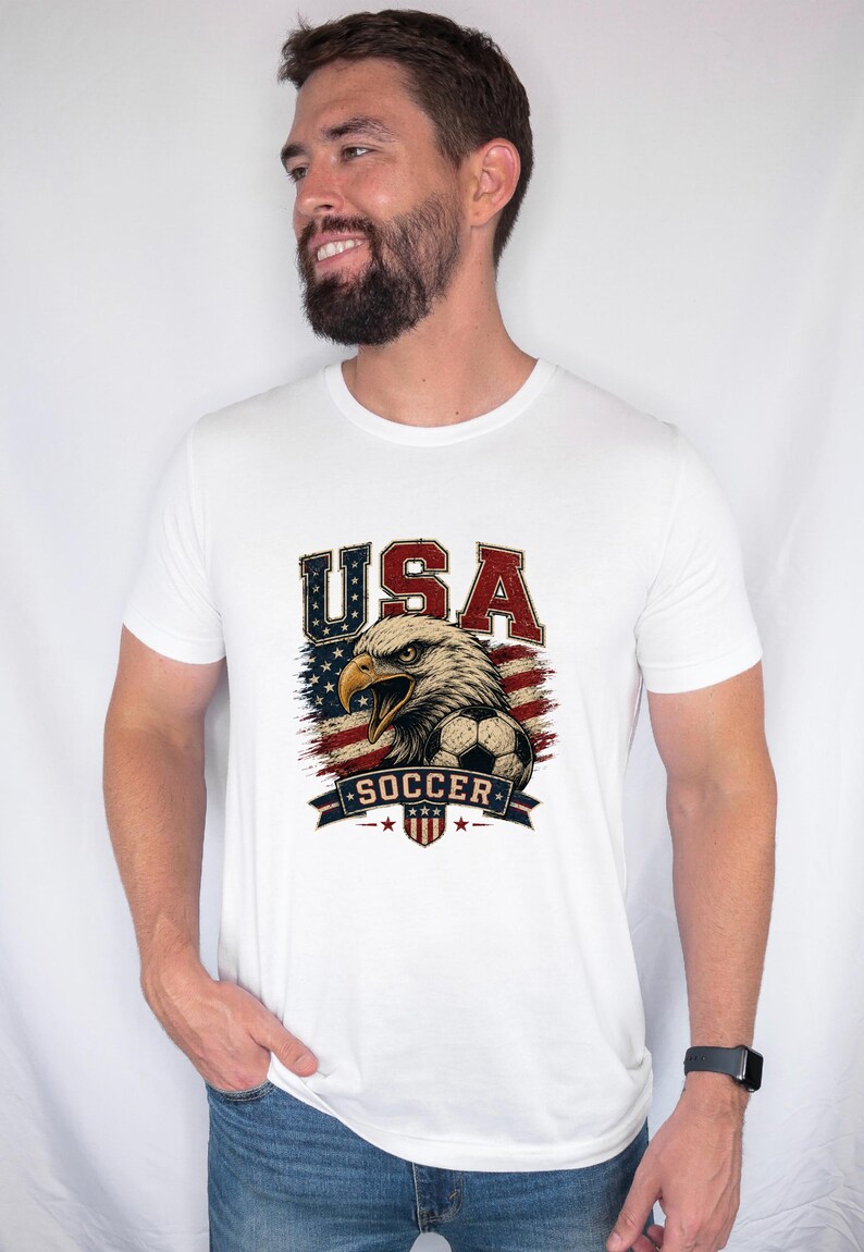 Puede incluir: Una camiseta blanca de manga corta con un estampado gr&aacute;fico de un &aacute;guila calva, un bal&oacute;n de f&uacute;tbol y la bandera estadounidense. El texto "USA" aparece en rojo y azul sobre el &aacute;guila, y la palabra "SOCCER" est&aacute; impresa en una banda azul debajo.