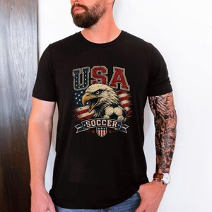 Puede incluir: Una camiseta negra de manga corta con un estampado gr&aacute;fico de un &aacute;guila calva, un bal&oacute;n de f&uacute;tbol y la bandera estadounidense. El texto "USA" aparece en rojo, blanco y azul sobre el &aacute;guila, con la palabra "SOCCER" impresa debajo del gr&aacute;fico.