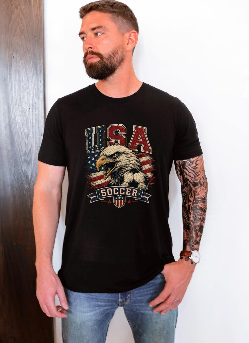 Puede incluir: Una camiseta negra de manga corta con un estampado gr&aacute;fico de un &aacute;guila calva, un bal&oacute;n de f&uacute;tbol y la bandera estadounidense. El texto "USA" aparece en rojo, blanco y azul sobre el &aacute;guila, con la palabra "SOCCER" impresa debajo del gr&aacute;fico.