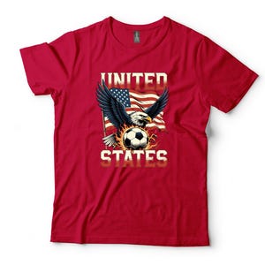 K&ouml;nnte beinhalten: Ein rotes Kurzarm-T-Shirt mit einer Grafik eines Wei&szlig;kopfseeadlers mit ausgebreiteten Fl&uuml;geln, einem Fu&szlig;ball und einer wehenden amerikanischen Flagge. Der Text UNITED STATES ist in einer fetten, hellen Schrift &uuml;ber und unter dem Motiv gedruckt.