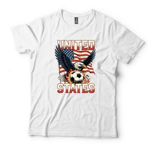 K&ouml;nnte beinhalten: Ein wei&szlig;es Kurzarm-T-Shirt mit einem Grafikdruck eines Wei&szlig;kopfseeadlers mit ausgebreiteten Fl&uuml;geln &uuml;ber einem von Flammen umgebenen Fu&szlig;ball. Dahinter ist eine wehende amerikanische Flagge zu sehen. Der Text UNITED STATES steht in roten Buchstaben.
