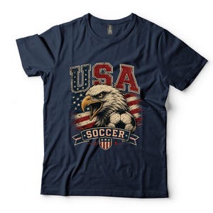Puede incluir: Una camiseta azul marino de manga corta con un estampado gr&aacute;fico de un &aacute;guila calva, un bal&oacute;n de f&uacute;tbol y la bandera estadounidense. El texto USA y SOCCER est&aacute; impreso en una fuente desgastada. El dise&ntilde;o incluye un peque&ntilde;o emblema de escudo.