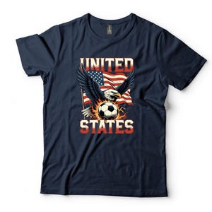 K&ouml;nnte beinhalten: Ein marineblaues Kurzarm-T-Shirt mit einem Grafikdruck eines Wei&szlig;kopfseeadlers mit ausgebreiteten Fl&uuml;geln, einem Fu&szlig;ball und einer amerikanischen Flagge. Der Text UNITED STATES ist in einer fetten, abgenutzten Schriftart aufgedruckt.