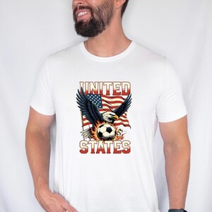K&ouml;nnte beinhalten: Ein wei&szlig;es Kurzarm-T-Shirt mit einem Grafikdruck eines Wei&szlig;kopfseeadlers mit ausgebreiteten Fl&uuml;geln &uuml;ber einem von Flammen umgebenen Fu&szlig;ball, vor dem Hintergrund einer US-Flagge mit dem Text UNITED STATES in fetten roten Buchstaben.