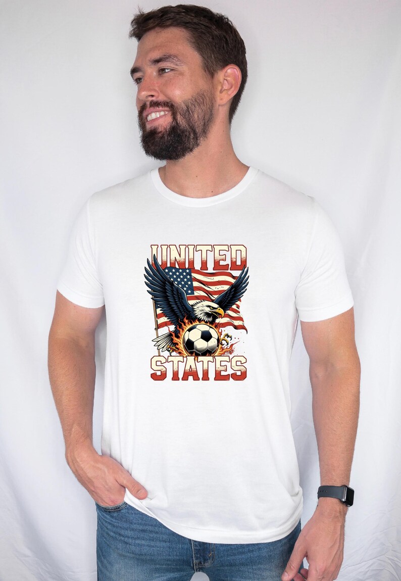K&ouml;nnte beinhalten: Ein wei&szlig;es Kurzarm-T-Shirt mit einem Grafikdruck eines Wei&szlig;kopfseeadlers mit ausgebreiteten Fl&uuml;geln &uuml;ber einem von Flammen umgebenen Fu&szlig;ball, vor dem Hintergrund einer US-Flagge mit dem Text UNITED STATES in fetten roten Buchstaben.