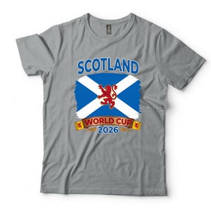 May include: Gray t-shirt featuring the word "SCOTLAND" above a blue and white flag design with a red lion emblem. Below the flag is "WORLD CUP 2026" in white text on a gold banner. A casual tee for fans.