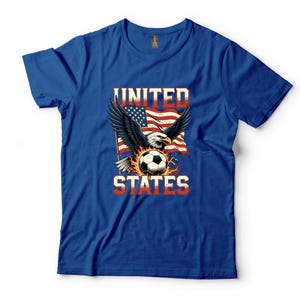 K&ouml;nnte beinhalten: Ein k&ouml;nigsblaues Kurzarm-T-Shirt mit einem Grafikdruck eines Wei&szlig;kopfseeadlers mit ausgebreiteten Fl&uuml;geln, einem von Flammen umgebenen Fu&szlig;ball und einer wehenden amerikanischen Flagge. Der Text UNITED STATES ist in fetten wei&szlig;en und roten Buchstaben aufgedruckt.