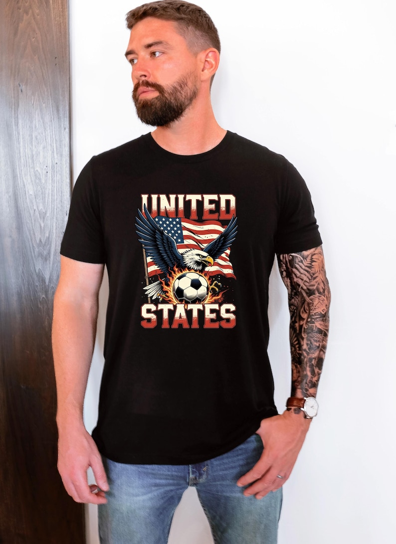 K&ouml;nnte beinhalten: Ein schwarzes Kurzarm-T-Shirt mit einem Grafikdruck eines Wei&szlig;kopfseeadlers mit ausgebreiteten Fl&uuml;geln &uuml;ber einem Fu&szlig;ball, vor einem stilisierten amerikanischen Flaggenmotiv. Der Text UNITED steht in Wei&szlig; dar&uuml;ber, STATES in Rot darunter.