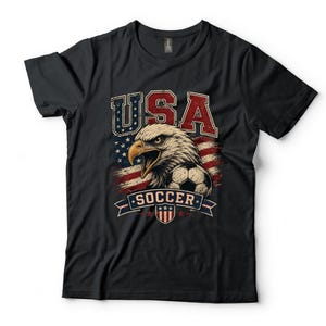 Puede incluir: Una camiseta negra de manga corta con un estampado gr&aacute;fico de un &aacute;guila calva, un bal&oacute;n de f&uacute;tbol y el texto USA SOCCER. El dise&ntilde;o incorpora colores rojo, blanco y azul con un patr&oacute;n de fondo de bandera estadounidense de estilo vintage.
