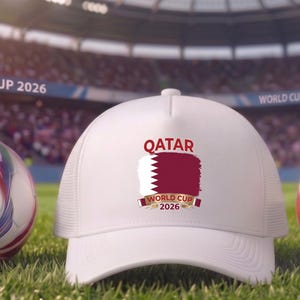 May include: A white baseball cap with the word "QATAR" and a maroon flag design, along with the text "WORLD CUP 2026". Two soccer balls with red, blue, and green accents are on either side of the cap, all on a grassy field.