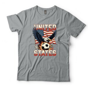 K&ouml;nnte beinhalten: Ein graues T-Shirt mit einer Grafik eines Wei&szlig;kopfseeadlers mit ausgebreiteten Fl&uuml;geln &uuml;ber einem von Flammen umgebenen Fu&szlig;ball. Hinter dem Adler ist eine wehende amerikanische Flagge zu sehen. Der Text UNITED STATES ist in fetten rot-wei&szlig;en Blockbuchstaben gedruckt.