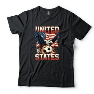 K&ouml;nnte beinhalten: Ein schwarzes Kurzarm-T-Shirt mit einem Grafikdruck eines Wei&szlig;kopfseeadlers mit ausgebreiteten Fl&uuml;geln &uuml;ber einem von Flammen umgebenen Fu&szlig;ball. Hinter dem Adler weht eine amerikanische Flagge. Der Text UNITED STATES ist in roten Buchstaben gedruckt.
