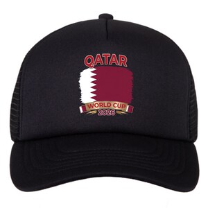 May include: Black trucker hat featuring the Qatar flag design with the text "QATAR WORLD CUP 2026". The hat has a mesh back and a curved brim. The Qatar flag is in maroon and white.