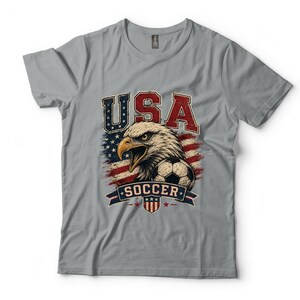 Puede incluir: Una camiseta gris de manga corta con un estampado gr&aacute;fico de un &aacute;guila calva, un bal&oacute;n de f&uacute;tbol y la bandera estadounidense. El texto USA est&aacute; impreso en rojo y azul sobre el &aacute;guila, y la palabra SOCCER aparece en una banda azul debajo.