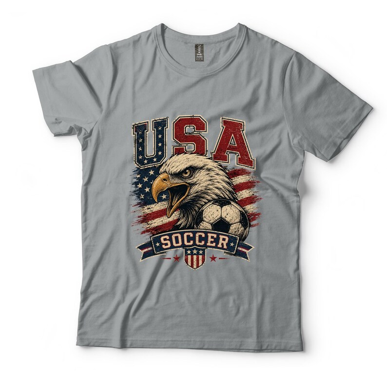 Puede incluir: Una camiseta gris de manga corta con un estampado gr&aacute;fico de un &aacute;guila calva, un bal&oacute;n de f&uacute;tbol y la bandera estadounidense. El texto USA est&aacute; impreso en rojo y azul sobre el &aacute;guila, y la palabra SOCCER aparece en una banda azul debajo.