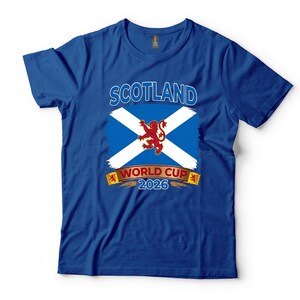 May include: Royal blue t-shirt featuring the Scotland flag with a white cross on a blue background and a red lion in the center. The text "SCOTLAND" is above the flag, and "WORLD CUP 2026" is below.