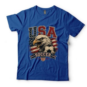 Puede incluir: Una camiseta de manga corta color azul real con un estampado gr&aacute;fico de un &aacute;guila calva, un bal&oacute;n de f&uacute;tbol y la bandera estadounidense. El texto USA aparece en rojo y blanco sobre el &aacute;guila, y la palabra SOCCER en blanco sobre una banda azul.