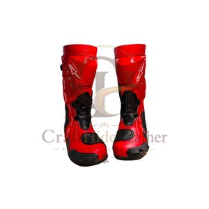 May include: Pair of red and black motorcycle boots. The boots have a tall design, with a red exterior and black accents on the toe, ankle, and shin areas. The boots feature a buckle and a white star logo.