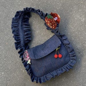 May include: A denim shoulder bag with a ruffled trim and strap. The bag features a flap closure and is adorned with a red strawberry charm and a pair of red cherry charms. A pink charm is attached to the side.
