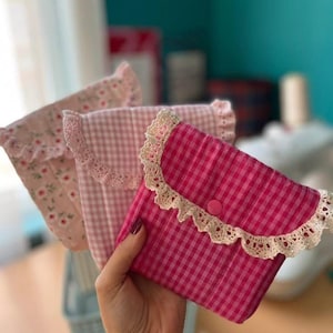 Può includere: Tre pochette in tessuto rosa. La pochette anteriore è un vivace rosa a quadretti con un bordo in pizzo bianco e una chiusura con bottone rosa. Le altre due pochette hanno motivi floreali e a quadretti, tutte con un bordo a balze.