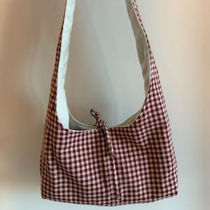 May include: A burgundy and cream gingham shoulder bag with a white interior. The bag has a long strap and a drawstring closure at the top. The bag is held up by a hand.