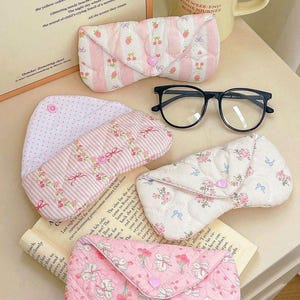 Könnte beinhalten: Eine Auswahl an gesteppten Brillenetuis in verschiedenen Pastellfarben, darunter Rosa und Weiß, mit Blumen- und Fruchtmustern. Eine Brille mit schwarzem Rahmen liegt daneben. Im Hintergrund ist ein beiger Becher mit Text zu sehen.
