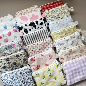 Puede incluir: Una colección de pequeñas bolsas rectangulares con cremallera en varios patrones. Los diseños incluyen motivos florales, de fresas, de vaca, de mariposas y de limones. Las bolsas son de tela y tienen una cremallera blanca.