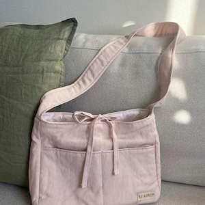 May include: A light pink shoulder bag with a long strap and two front pockets. The bag has a drawstring closure and a small label that reads "by ayarn". The bag is made of a soft, textured fabric.