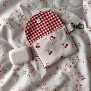 Op de afbeelding: Een witte AirPod-case naast een stoffen etui. De etui heeft een rood-witte ruitjes binnenkant en een witte buitenkant met een kersenpatroon. Het heeft een zilveren sluiting en een witte drukknoopsluiting.