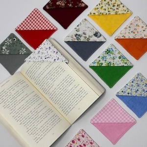 Puede incluir: Una colección de marcadores de libros de tela hechos a mano en varios patrones y colores, incluyendo florales, cuadros y tonos lisos. Los marcadores de libros en forma de diamante están colocados alrededor de un libro abierto, mostrando su uso.