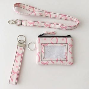 Könnte beinhalten: Ein passendes Accessoire-Set aus hellrosa Stoff mit einem sich wiederholenden Muster aus rosa Schleifen und Herzen. Das Set enthält ein Schlüsselband mit Silberverschluss, einen Schlüsselanhänger und eine kleine Reißverschlusstasche mit transparentem Ausweisfenster.