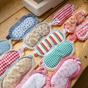 May include: A collection of handmade sleep masks in various patterns and colors, including floral, gingham, and striped designs. Each mask features a soft fabric and a lace trim, with a ribbon tie. The masks are arranged on a wooden surface near a sewing machine.