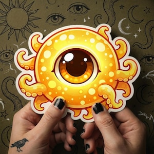 May include: A yellow and orange sticker featuring a cartoon eye with tentacles. The eye has a brown iris and a yellow pupil. The sticker is held by two hands with black nail polish and a crow tattoo.