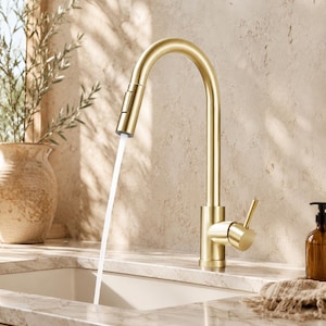 May include: A gold-colored kitchen faucet with a high arc spout. Water flows from the faucet into a white sink. The faucet has a single lever handle and a pull-down sprayer. The background includes a vase with greenery.