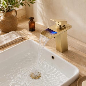 May include: A contemporary gold-coloured bathroom tap with a rectangular spout, featuring blue illumination, dispensing water into a white rectangular sink. The tap is set on a beige countertop, with a vase and a small bottle in the background.