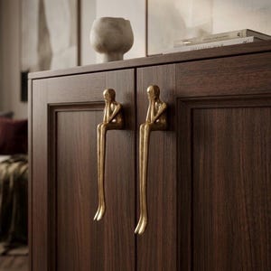 Puede incluir: Un armario de madera marrón oscuro con dos tiradores dorados. Cada tirador es una figura estilizada de una persona sentada con la barbilla apoyada en la mano. El armario tiene un diseño sencillo y elegante.