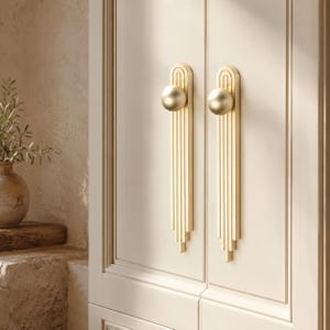 May include: Two vertical gold-toned metal cabinet handles mounted on a light-colored door. Each handle features a smooth spherical knob at the top, positioned above a series of parallel vertical bars that extend downward in a stepped design.