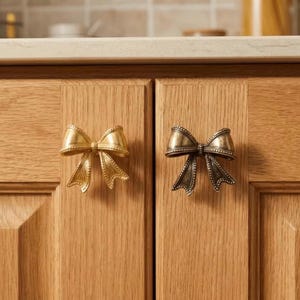 Peut inclure: Deux poignées de placard en forme de nœud papillon, une dorée et une bronze, montées sur une porte de placard en bois clair. Ces poignées décoratives ajoutent une touche d'élégance à la menuiserie.