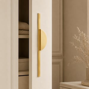 Peut inclure: Une poignée de placard verticale en métal doré composée d'une longue barre rectangulaire fine avec une prise en demi-cercle fixée au centre. La quincaillerie est montée sur une porte de placard claire au fini lisse et mat.