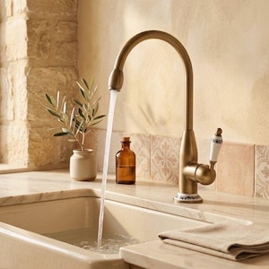 May include: A bronze kitchen tap with a high arched spout, water flowing into a white sink. The tap has a white handle with a decorative design. A small brown bottle and a potted plant are visible.