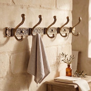 Peut inclure: Un porte-manteau mural décoratif de couleur bronze avec cinq crochets incurvés. Le porte-manteau présente des détails floraux ornés et une serviette gris clair suspendue à un crochet. Le porte-manteau est fixé sur un mur de briques de couleur claire.