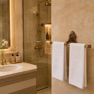 Peut inclure: Scène de salle de bain avec un porte-serviettes doré tenant deux serviettes blanches rectangulaires. Le porte-serviettes a un design orné. L'arrière-plan comprend une douche avec des accessoires dorés et un comptoir en marbre avec un évier.