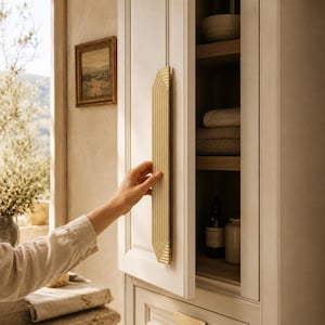 Peut inclure: Une longue poignée de placard verticale en or brossé, dotée d'une série de crêtes horizontales parallèles en relief sur toute sa longueur. La poignée est fixée sur une porte de placard crème entrouverte, révélant des étagères en bois avec du linge.