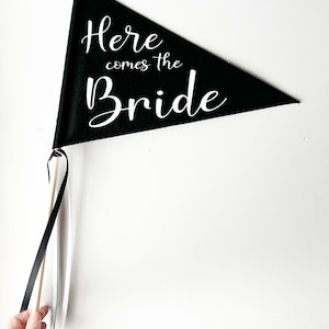 May include: Black triangular flag with white text that reads "Here comes the Bride". The flag is attached to a wooden stick with black and white ribbon streamers.