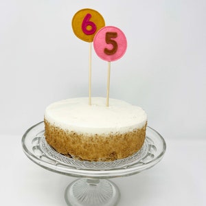 May include: A white cake with brown sprinkles on a glass cake stand. The cake is decorated with two felt numbers, a pink 5 and a gold 6.