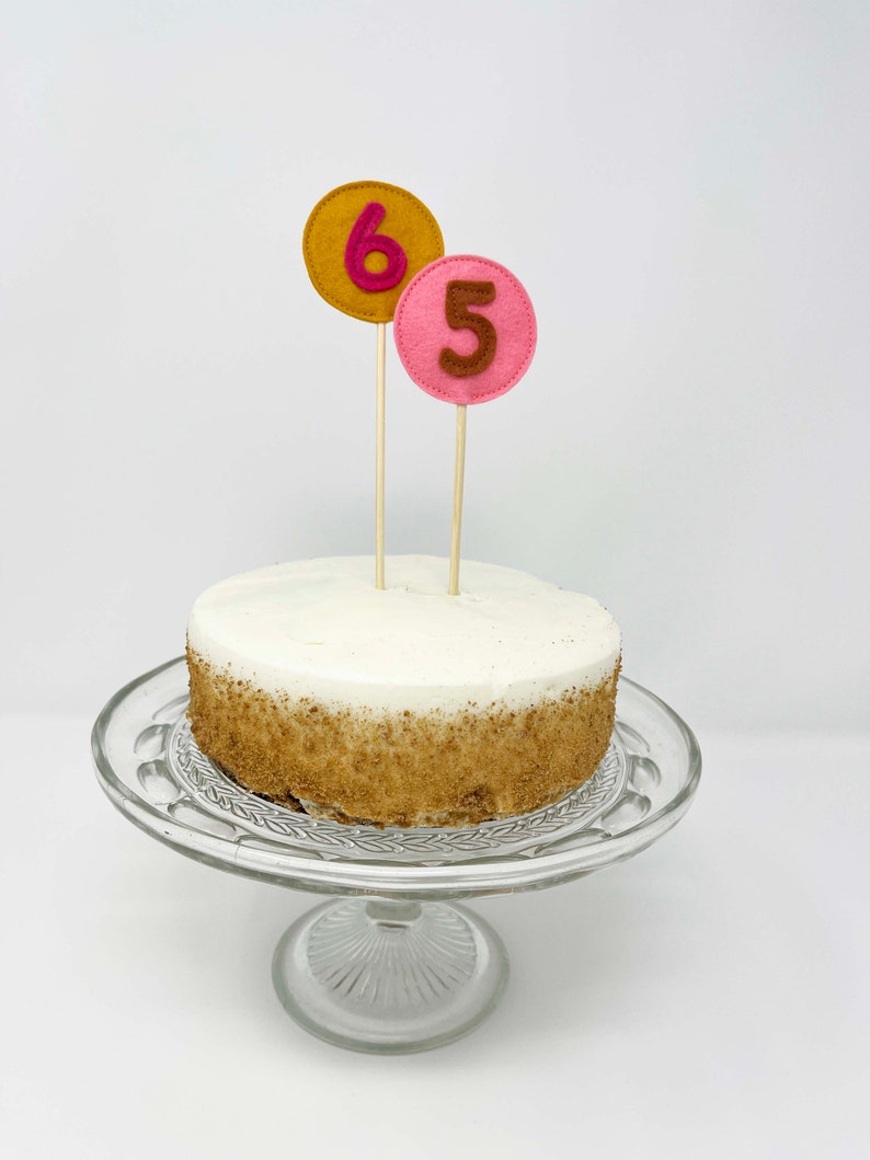 May include: A white cake with brown sprinkles on a glass cake stand. The cake is decorated with two felt numbers, a pink 5 and a gold 6.