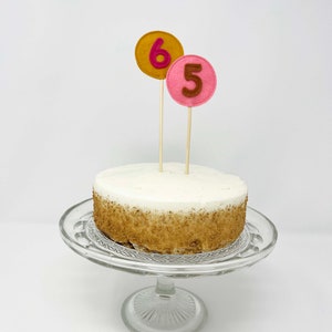 May include: A white cake with brown sprinkles on a glass cake stand. The cake is decorated with two felt numbers, a pink "5" and a yellow "6".