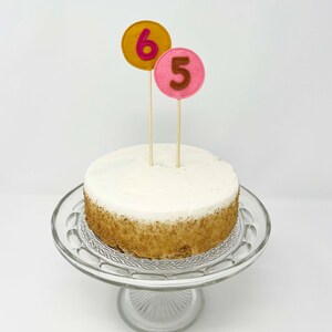 May include: A white cake with brown crumbs on the bottom tier, decorated with two felt numbers, a pink "5" and a yellow "6", on wooden sticks.
