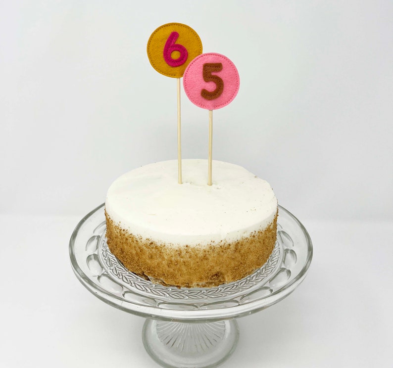 May include: A white cake with brown crumbs on the bottom tier, decorated with two felt numbers, a pink "5" and a yellow "6", on wooden sticks.